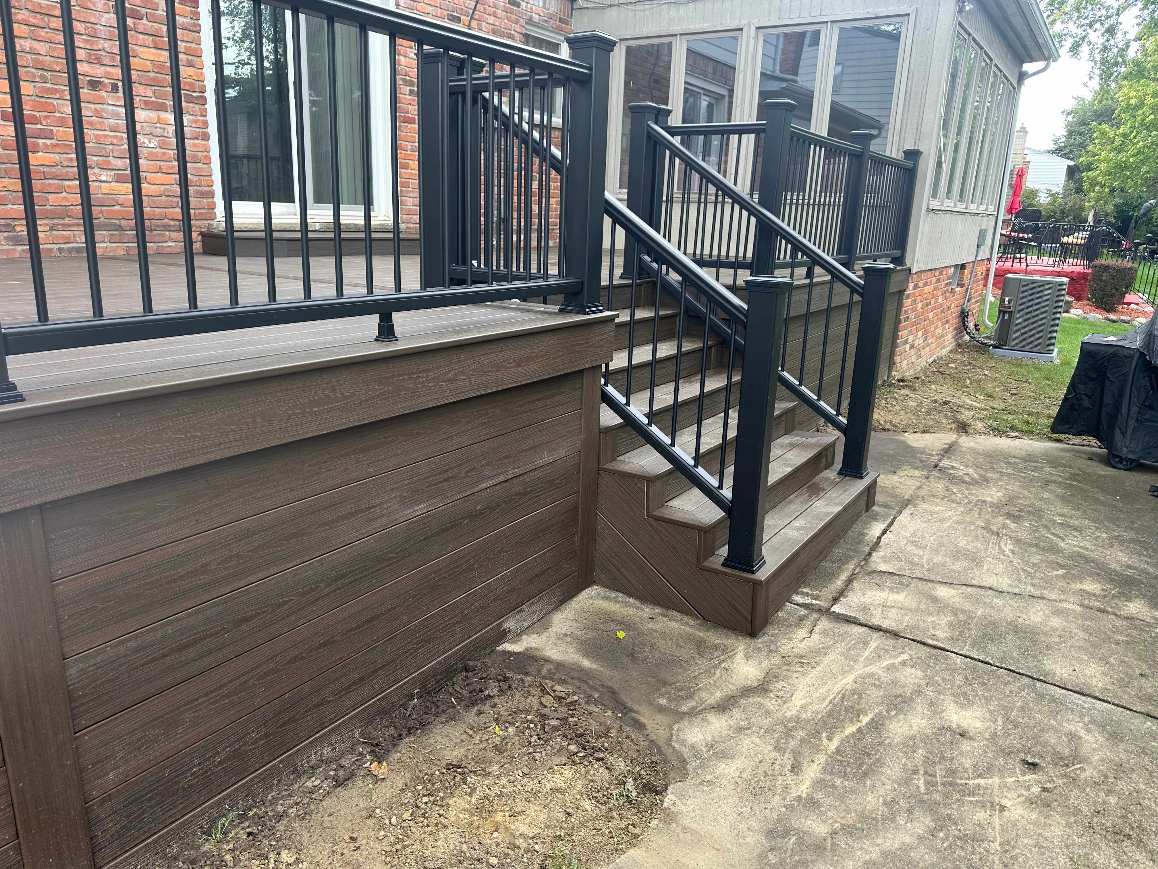 Multi-level composite deck with glass and black metal railings, stairs, and brick home integration